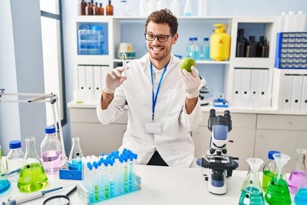 Young Hispanic Man Working Scientist Laboratory Holding Apple Winking Looking — Stok fotoğraf