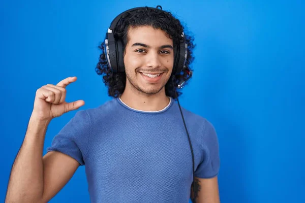 ヘッドフォンを使用して音楽に耳を傾け巻き毛を持つヒスパニック系の男は笑顔と指を見てとカメラと小さなサイズの看板を行う手で自信を持ってジェスチャー 概念を測定し — ストック写真