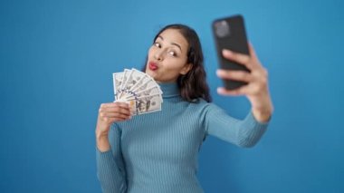 Genç ve güzel İspanyol kadın, izole edilmiş mavi arka planda dolarları tutarak selfie çekiyor.