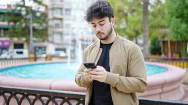 Genç İspanyol adam parkta gülümseyen akıllı telefon kullanıyor.