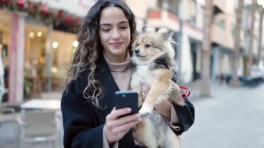 Köpeğiyle gülümseyen genç İspanyol kadın. Sokakta akıllı telefon kullanıyor.