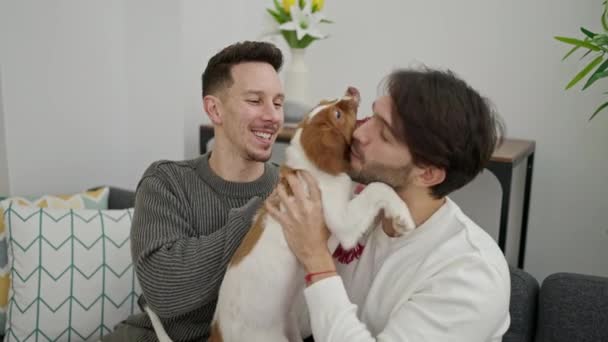 Dwóch Mężczyzn Uśmiecha Się Pewnie Siedząc Kanapie Całując Psa Domu — Wideo stockowe