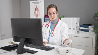 Genç beyaz kadın doktor klinikte telefonla konuşurken bilgisayar kullanıyor.