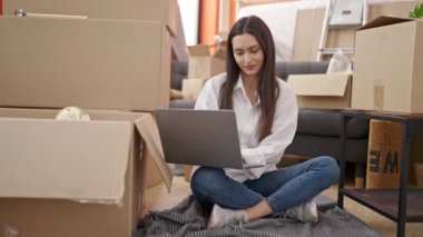 Young beautiful hispanic woman using laptop sitting on floor at new home
