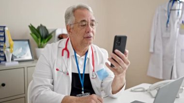 Gri saçlı bir doktorla orta yaşlı bir adam klinikte akıllı telefonuyla video görüşmesi yapıyor.