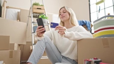 Genç sarışın kadın yeni evinde akıllı telefon ve kredi kartıyla alışveriş yapıyor.
