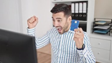 Genç İspanyol iş adamı kredi kartıyla alışveriş yapıyor ve ofisteki kazanan ifadeyle bilgisayar kullanıyor.