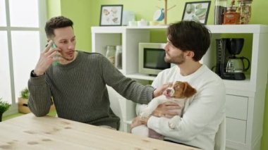 İki erkek akıllı telefondan konuşuyor. Yemek salonunda köpekle masada oturuyorlar.