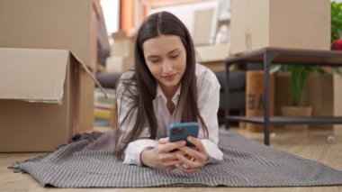 Genç, güzel, İspanyol bir kadın yeni evinde yerde yatarken akıllı telefon kullanıyor.
