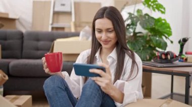 Genç ve güzel İspanyol kadın akıllı telefon kullanıyor. Yeni evinde yerde kahve içiyor.