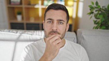 Kanepede oturan Latin asıllı genç adam evde çenesini kapatıp jest yapıyor.