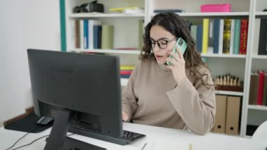 Young beautiful hispanic woman student using computer talking on smartphone at library university