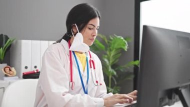 Klinikte bilgisayar kullanarak tıbbi maske takan genç, güzel İspanyol kadın doktor.