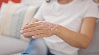 Orta yaşlı İspanyol bir kadın, evinde koltukta oturup cilt bakımı yapıyor.