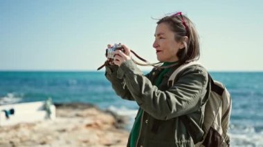 Sırt çantası takan gri saçlı, olgun İspanyol bir kadın sahilde fotoğraf makinesi ile fotoğraf çekiyor.