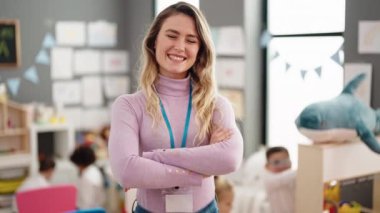 Young blonde woman teacher smiling confident standing with arms crossed gesture at kindergarten