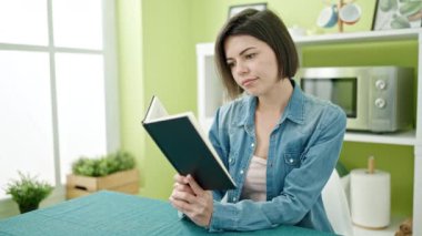 Genç beyaz kadın kitap okuyor. Evdeki masada oturuyor.