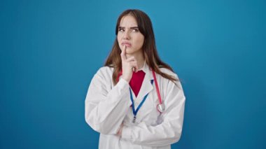 Şüpheli bir ifadeyle duran genç İspanyol kadın doktor, izole edilmiş mavi arka plan üzerinde fikir yürütüyor.