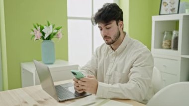 Genç İspanyol adam yemek odasındaki masada dizüstü bilgisayar ve akıllı telefon kullanıyor.
