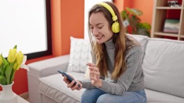 Genç beyaz kadın evde kanepede oturmuş müzik dinliyor.