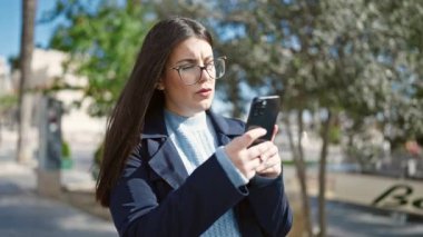 Genç İspanyol kadın parkta akıllı telefon kullanarak gülümsüyor.