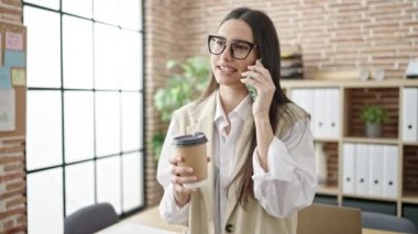 Young beautiful hispanic woman business worker talking on smartphone drinking coffee at office