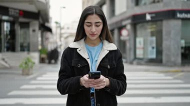 Genç, güzel, İspanyol bir kadın sokakta akıllı telefon kullanarak gülümsüyor.