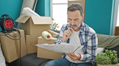 Orta yaşlı bir adam yeni evindeki kanepede oturmuş belge okuyor.