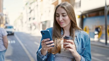 Young beautiful hispanic woman using smartphone drinking coffee at street