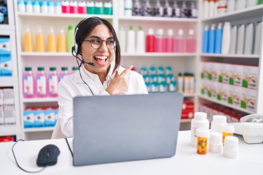 Eczanede çalışan genç bir Arap kadın yüzünde kocaman bir gülümsemeyle dizüstü bilgisayar kullanarak kameraya el ve parmak ile bakıyor.. 