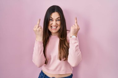 Young brunette woman standing over pink background showing middle finger doing fuck you bad expression, provocation and rude attitude. screaming excited 