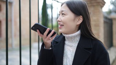 Young chinese woman smiling confident sending voice message by smartphone at street