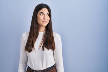 Genç esmer kadın mavi arka planda duruyor gülümsüyor, yan tarafa bakıyor ve düşünüyor.. 