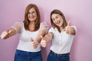 İspanyol anne ve kızı pembe arka plan üzerine gündelik beyaz tişört giyip olumlu jestler yapıyorlar. Elleriyle gülümsüyorlar ve başarı için mutlular. kazanan jest. 