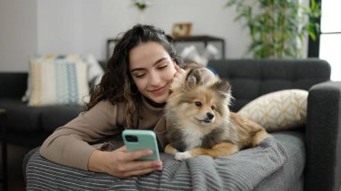 Köpekli genç İspanyol kadın evdeki kanepede uzanmış akıllı telefon kullanıyor.