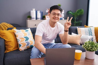 Genç Asyalı adam evinde dizüstü bilgisayar kullanıyor kanepede oturuyor gülümsüyor ve parmaklarını zafer işareti yaparken kameraya bakıyor. İki numara.. 