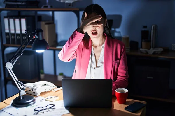 Chinese young woman working at the office at night peeking in shock covering face and eyes with hand, looking through fingers with embarrassed expression.