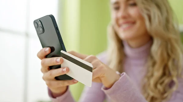 年轻的金发女人拿着智能手机和信用卡坐在饭厅的桌子上购物 — 图库照片
