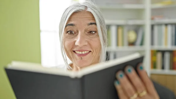 Middle age grey-haired woman student reading book smiling at library university