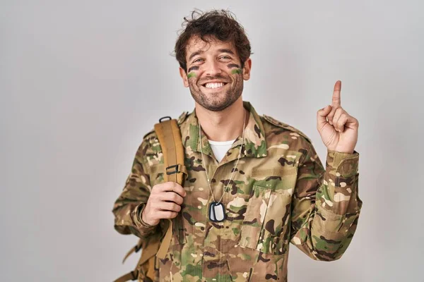 顔に大きな笑顔でカモフラージュ軍の制服を着たヒスパニック系の若い男 カメラを見て側に手指で指して — ストック写真