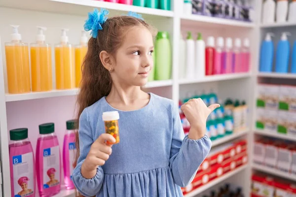 Young Little Girl Holding Pills Pharmacy Pointing Thumb Side Smiling — ストック写真
