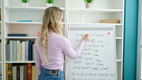 Young blonde woman teacher teaching maths lesson writing on white board at classroom