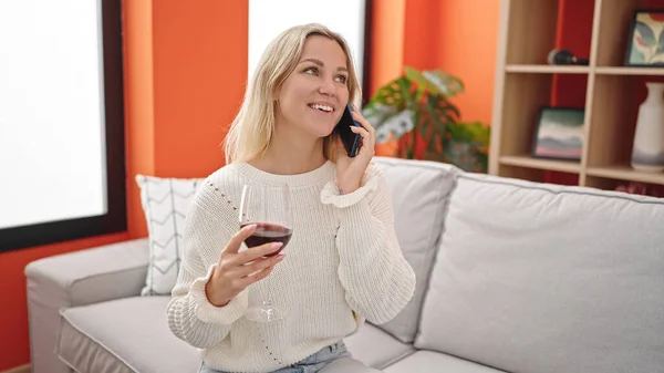 Mujer Rubia Joven Hablando Teléfono Inteligente Bebiendo Vino Casa Imagen de archivo