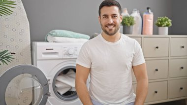 Çamaşır odasında yerde oturmuş kendinden emin gülümseyen İspanyol genç adam.