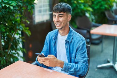 Young hispanic man using smartphone sitting on table at coffee shop terrace