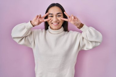 Pembe arka planda duran genç Güney Asyalı kadın barış sembolü yapıyor. Yüzünde parmakları, gülümseyerek zaferi gösteriyor. 