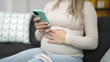 Genç hamile kadın akıllı telefon kullanıyor. Evde karnına dokunuyor.
