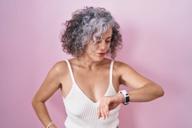 Pembe arka planda gri saçlı orta yaşlı bir kadın kol saatini kontrol ediyor, rahat ve kendinden emin. 