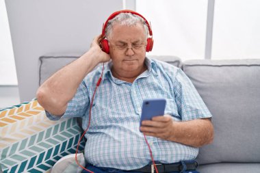 Orta yaşlı, kır saçlı, müzik dinleyen adam evdeki kanepede oturuyor.