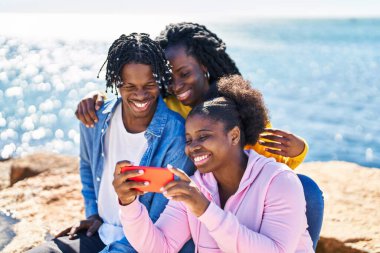African american friends using smartphone sitting on rock at seaside